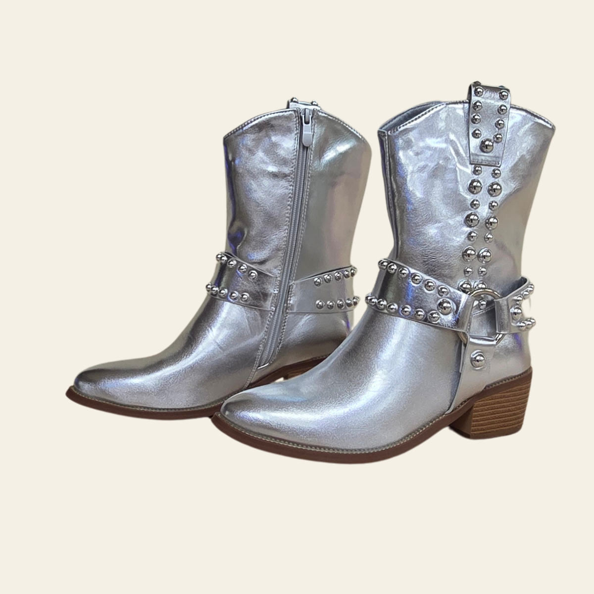 A pair of metallic silver western-style boots with pointed toes, brown block heels, and silver studded straps with buckles, shown against a solid light beige background.