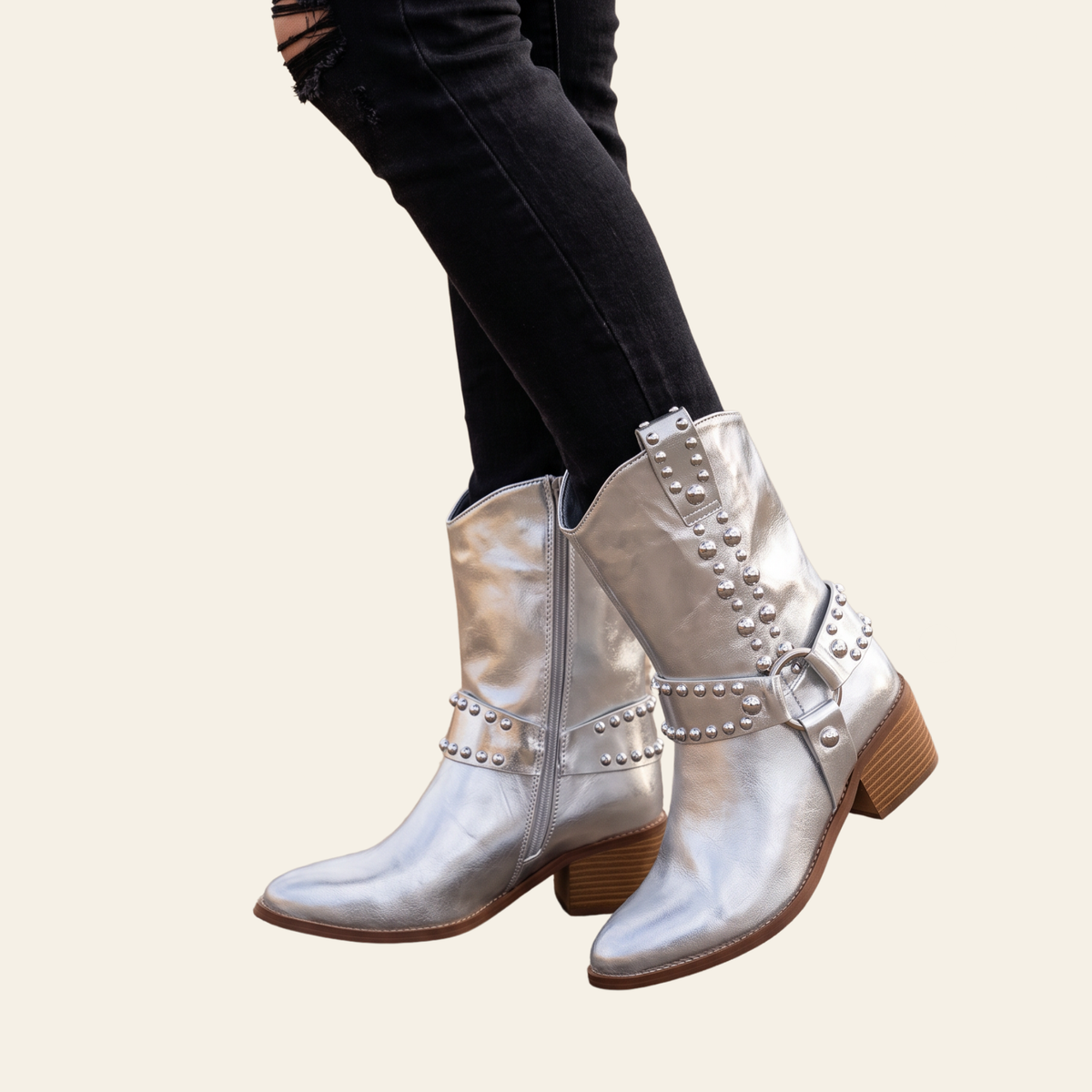 A pair of metallic silver western-style ankle boots with studded straps and brown block heels, shown on a model wearing black ripped jeans against a light beige background.