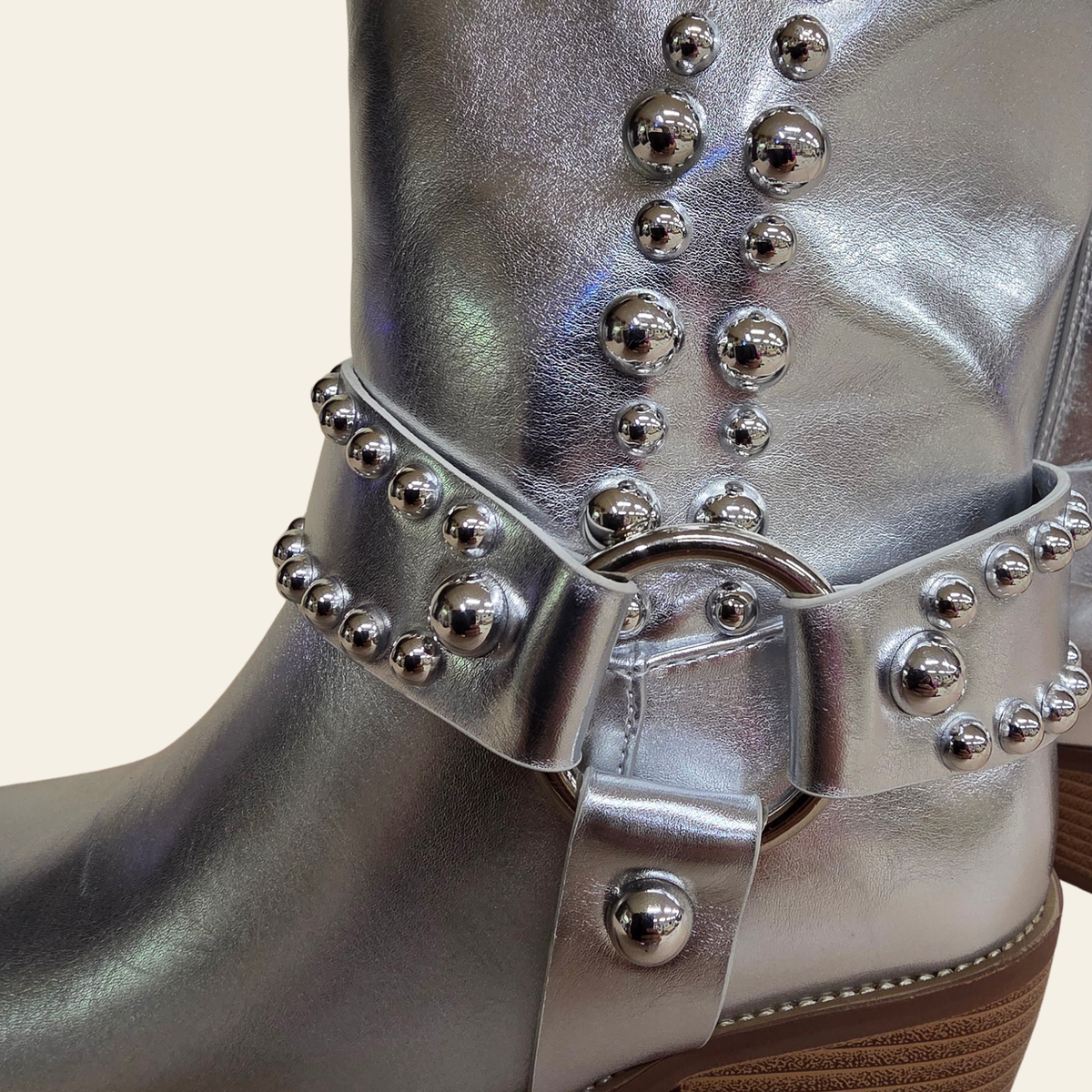 A close-up of a metallic silver studded boot with an ankle strap and buckle, set against a plain light beige background.