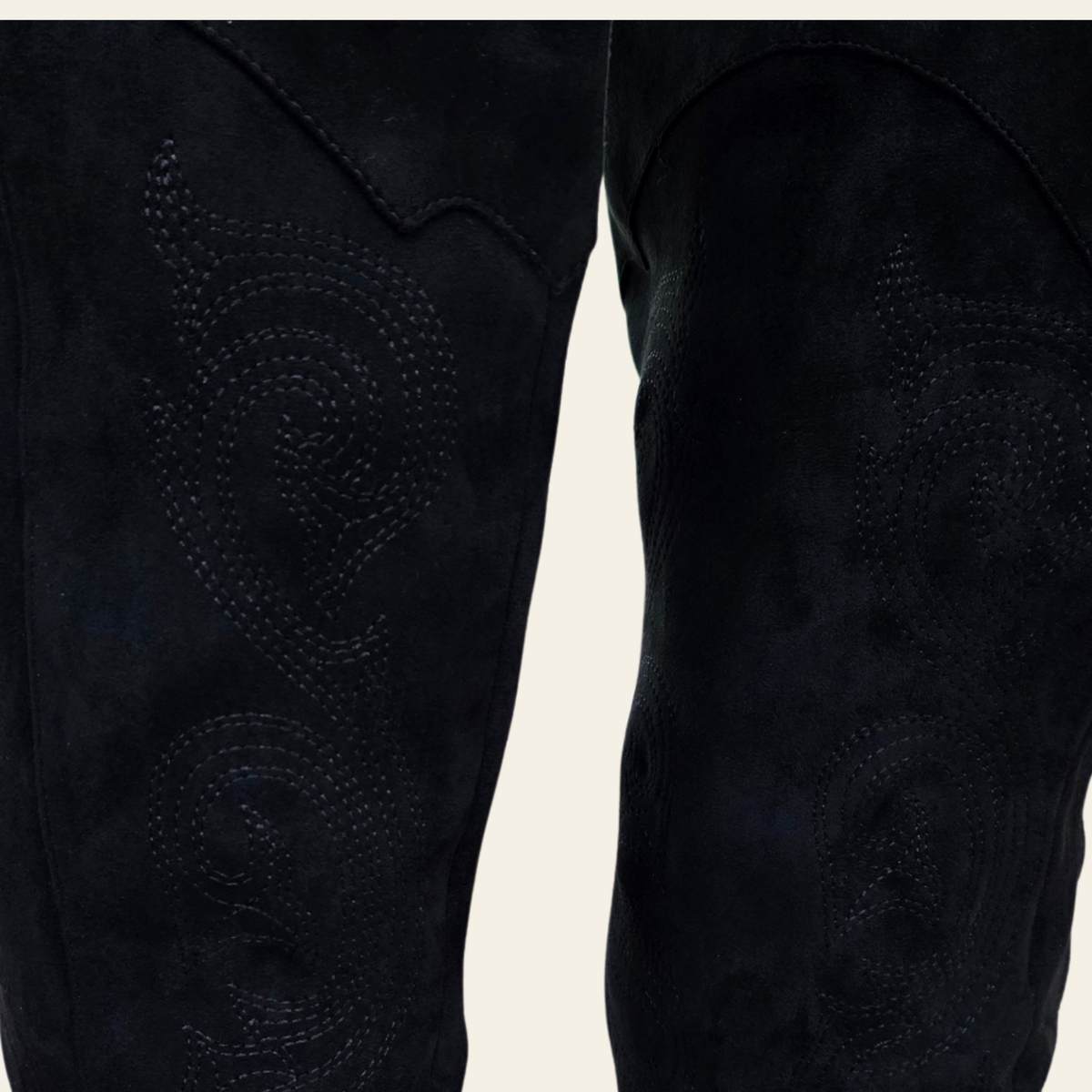 Close-up of black suede western-style pants showing detailed embroidered stitching on the upper legs against a light off-white background.