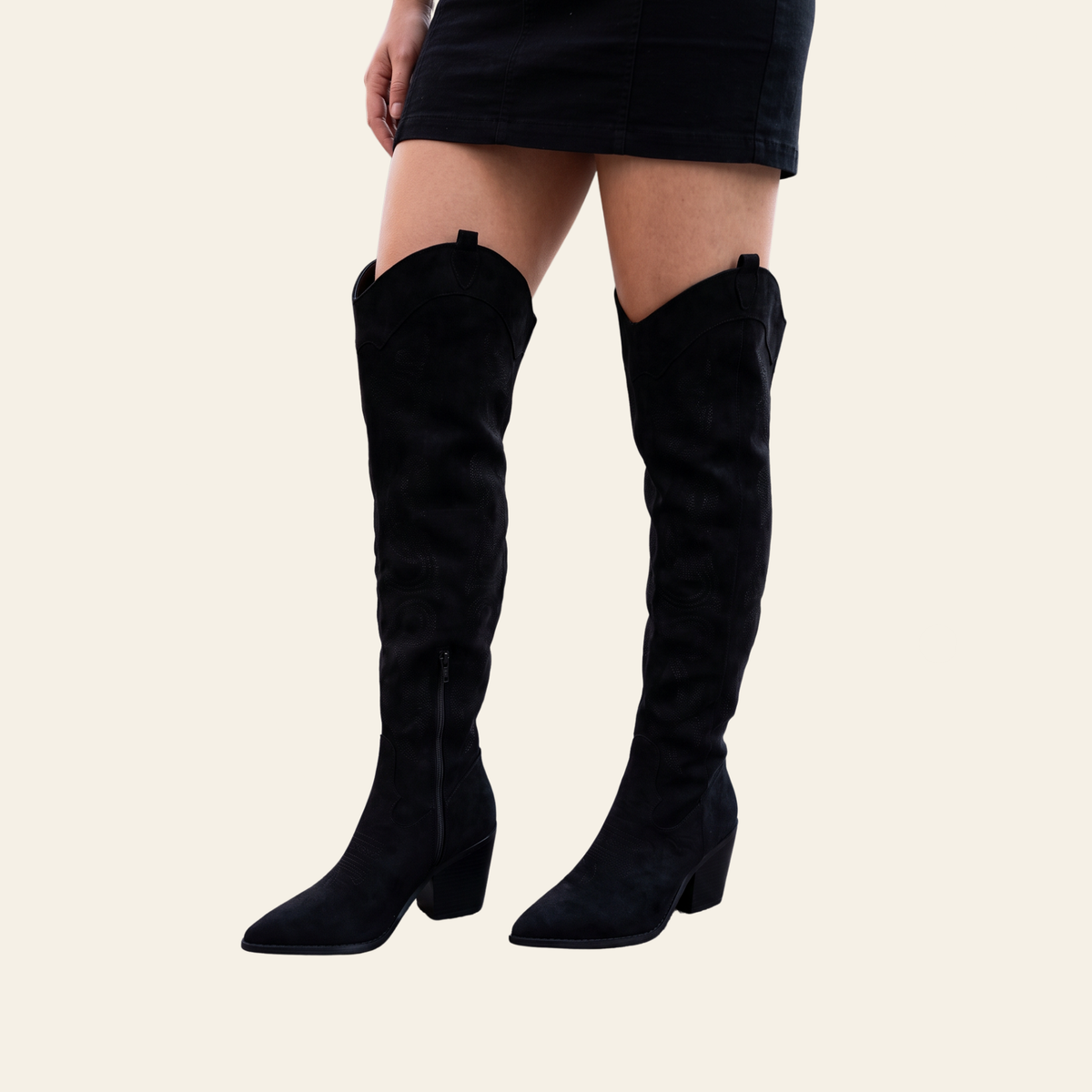 A person wearing black suede over-the-knee boots with a block heel, shown from the waist down against a plain light beige background.