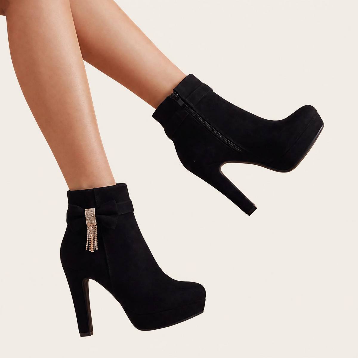 Black suede ankle boots with platform soles, high stiletto heels, and a decorative rhinestone bow with dangling fringe on one boot, shown on a model's feet against a light beige background.