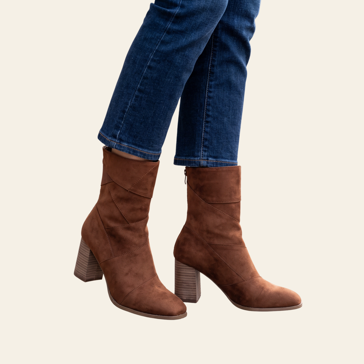 A pair of brown suede ankle boots with block heels, shown on a model wearing blue jeans against a light beige background.