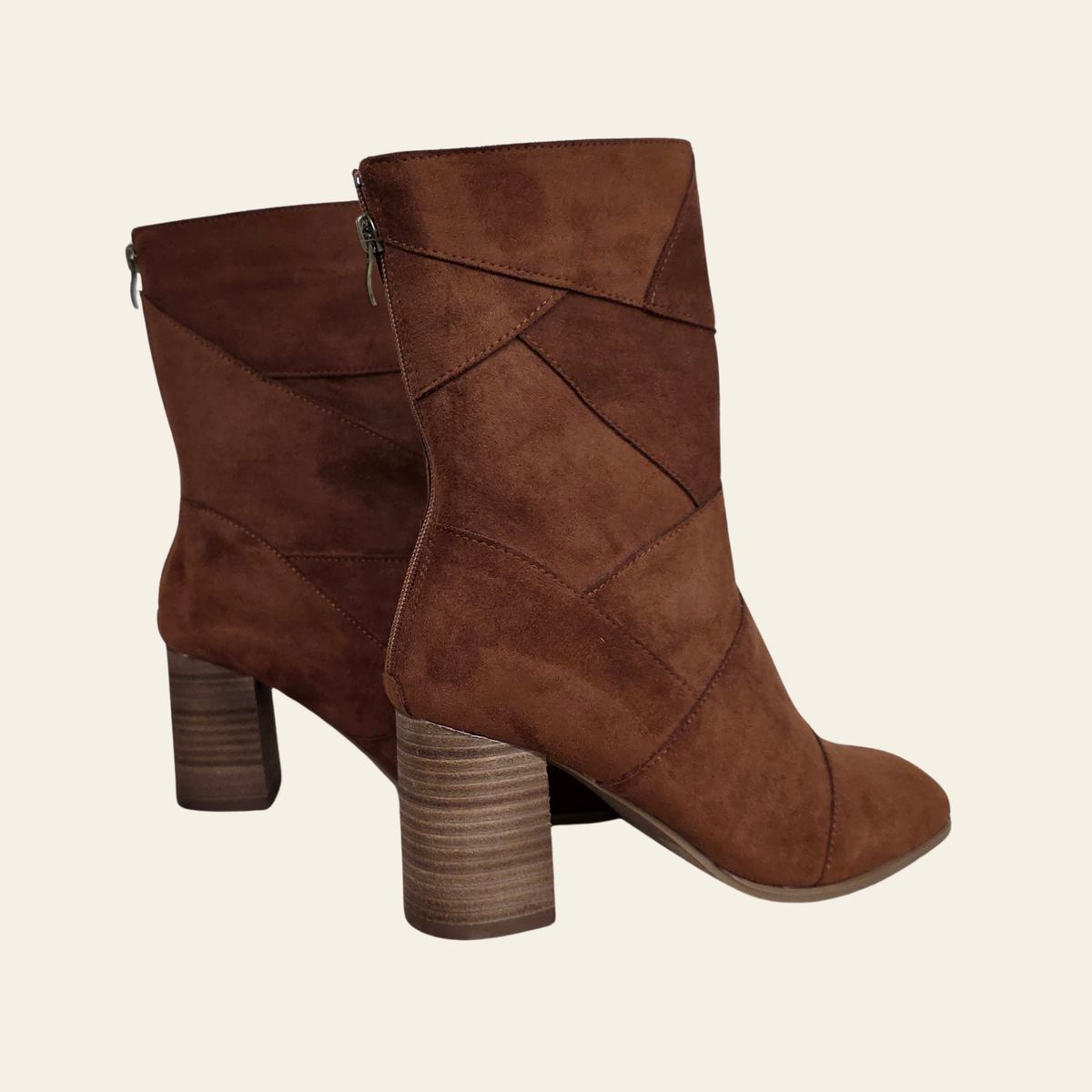 A pair of brown suede ankle boots with a stacked block heel and geometric paneling on the upper, shown against a solid off-white background.