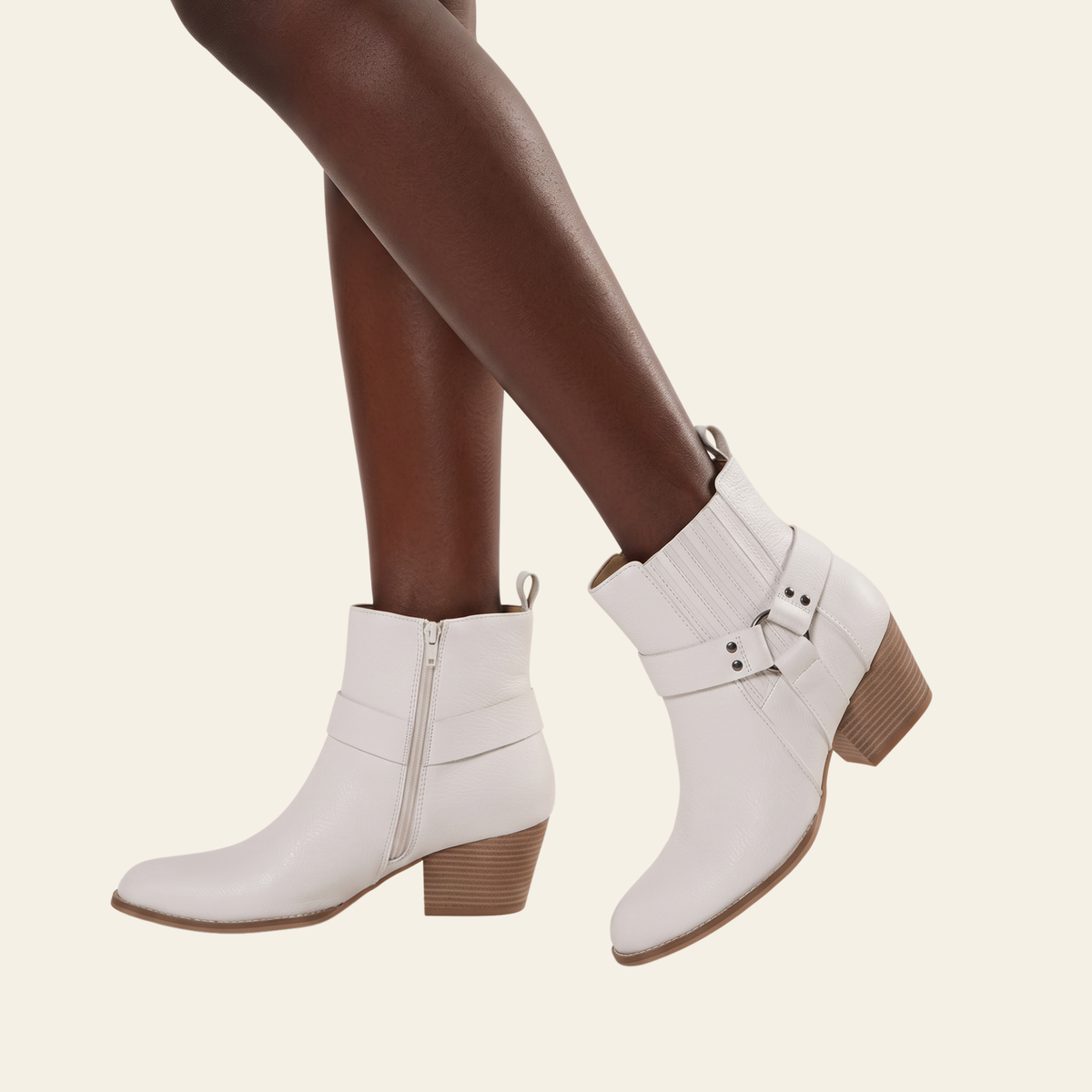 White vegan leather ankle boots with a side zipper and decorative buckle strap, shown on a model's feet against a light beige background.