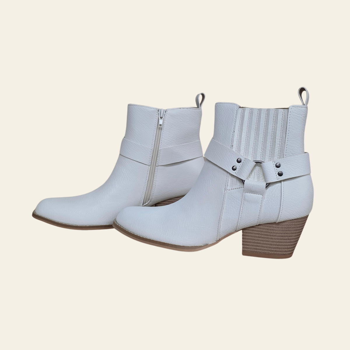 A pair of white faux leather ankle boots with side zippers, decorative straps, and stacked block heels, shown against a solid light beige background.