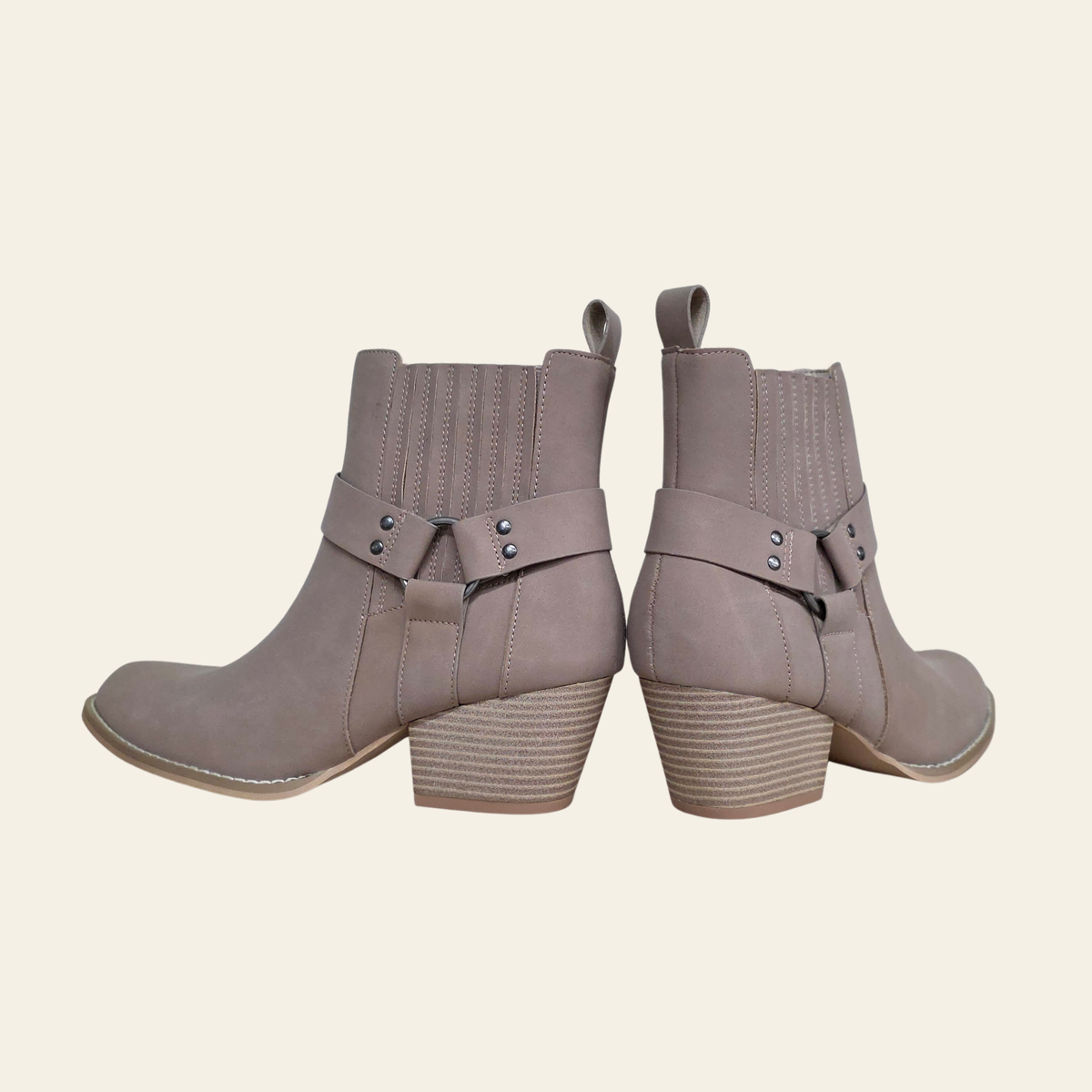 A pair of taupe-brown ankle boots with a western-inspired design, featuring a stacked block heel, a decorative strap with silver-tone studs across the instep, and a pull tab at the back collar, shown from a side-rear angle on a light cream background.