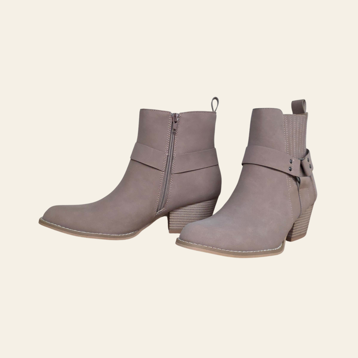 A pair of taupe-brown ankle boots with side zippers and decorative straps, shown against a solid cream background.
