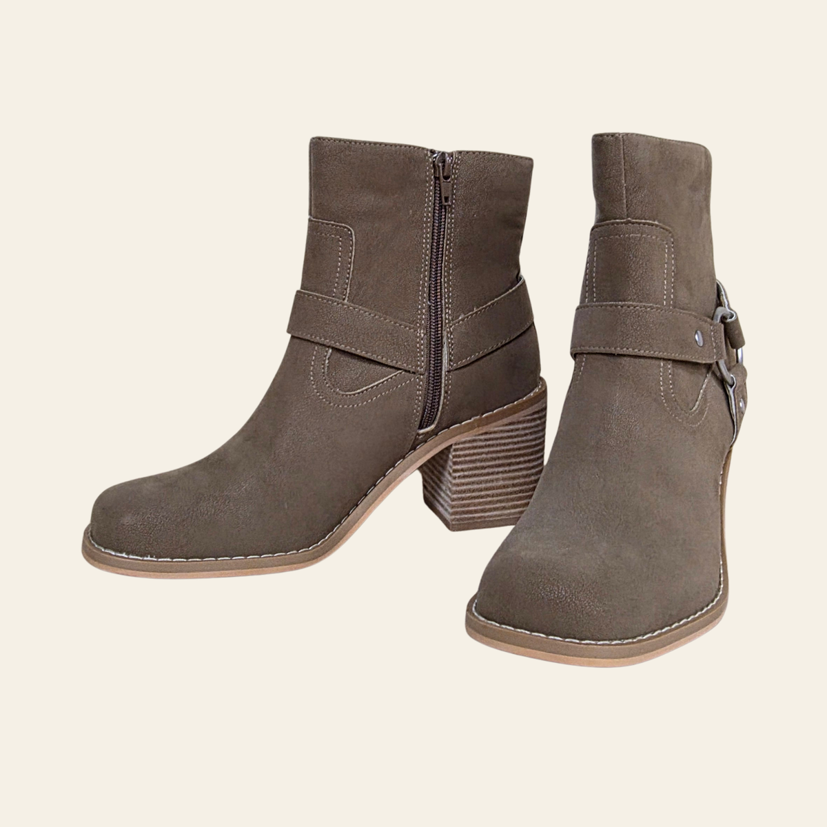 A pair of taupe-brown suede ankle boots with side zippers, wrap-around strap details, and stacked block heels, shown against a solid cream background.