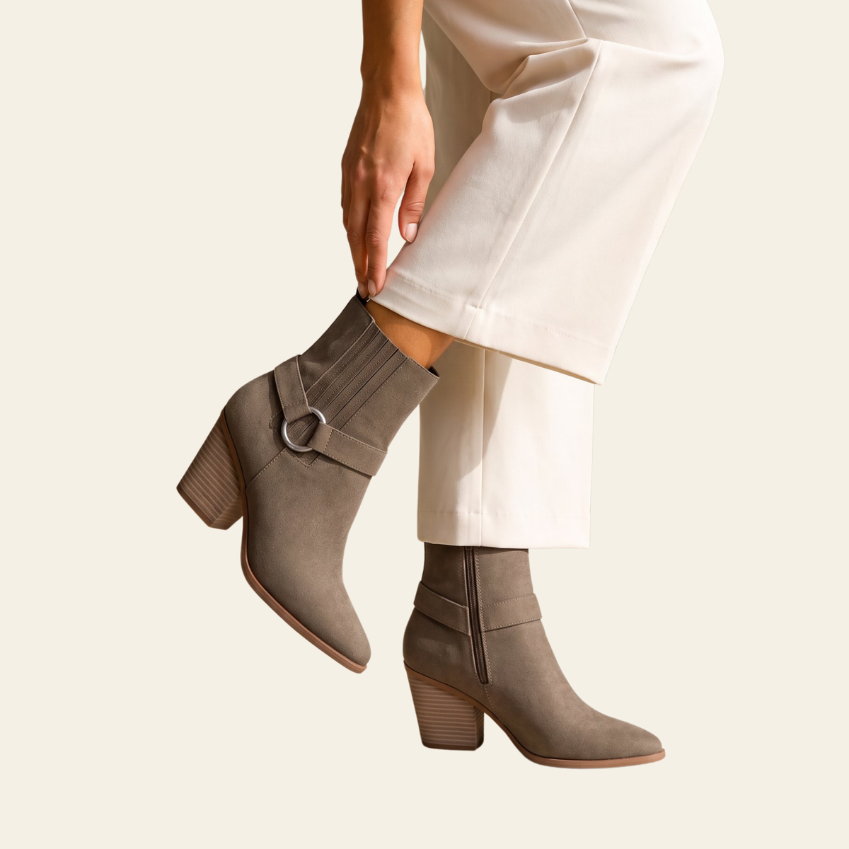 A person wearing taupe-brown ankle boots with a side buckle strap and block heel, paired with off-white cropped pants, against a solid light beige background.