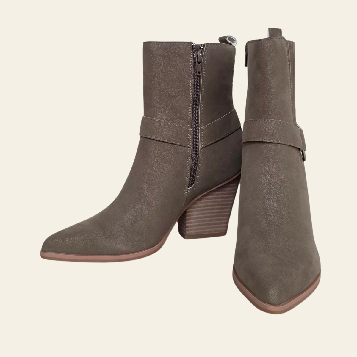 A pair of taupe-brown suede ankle boots with side zippers, block heels, and a decorative strap detail on the side, shown against a solid cream background.