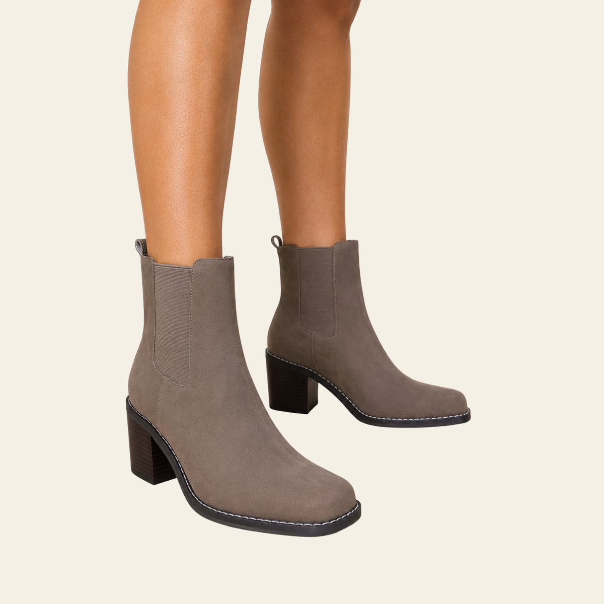 A pair of taupe-brown faux suede Chelsea-style ankle boots with block heels, shown on a model's feet against a solid cream background.