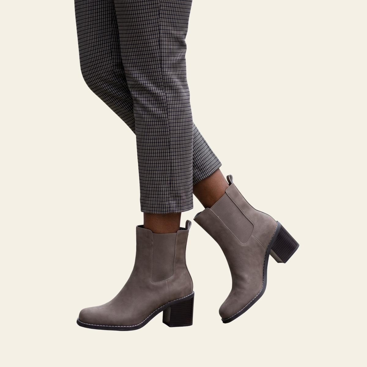 A pair of taupe gray faux suede Chelsea-style ankle boots with block heels, shown on a model wearing gray and black checkered trousers against a light cream background.