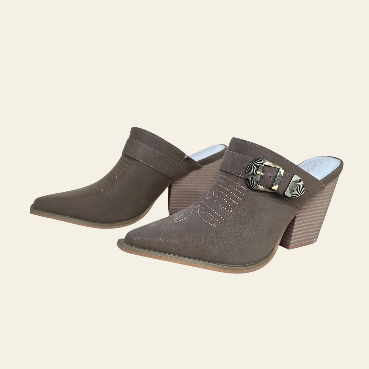 A pair of women's pointed-toe mule heels in taupe brown suede, featuring a wide strap with a silver-tone buckle and decorative stitching on the vamp, set against a solid cream background.