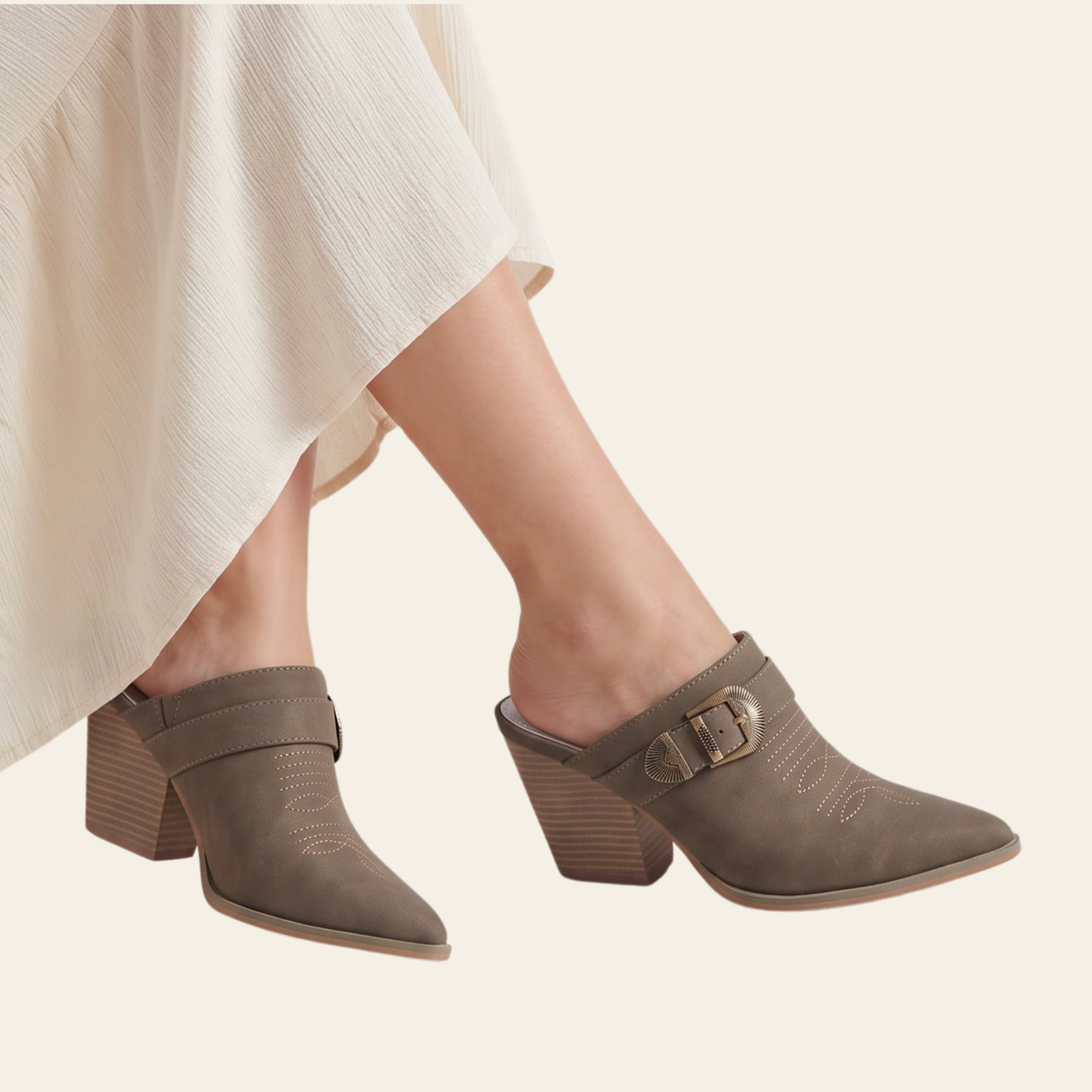 A pair of taupe-brown mule heels with block toes and buckle straps, shown on a model wearing a cream-colored skirt against a solid light beige background.