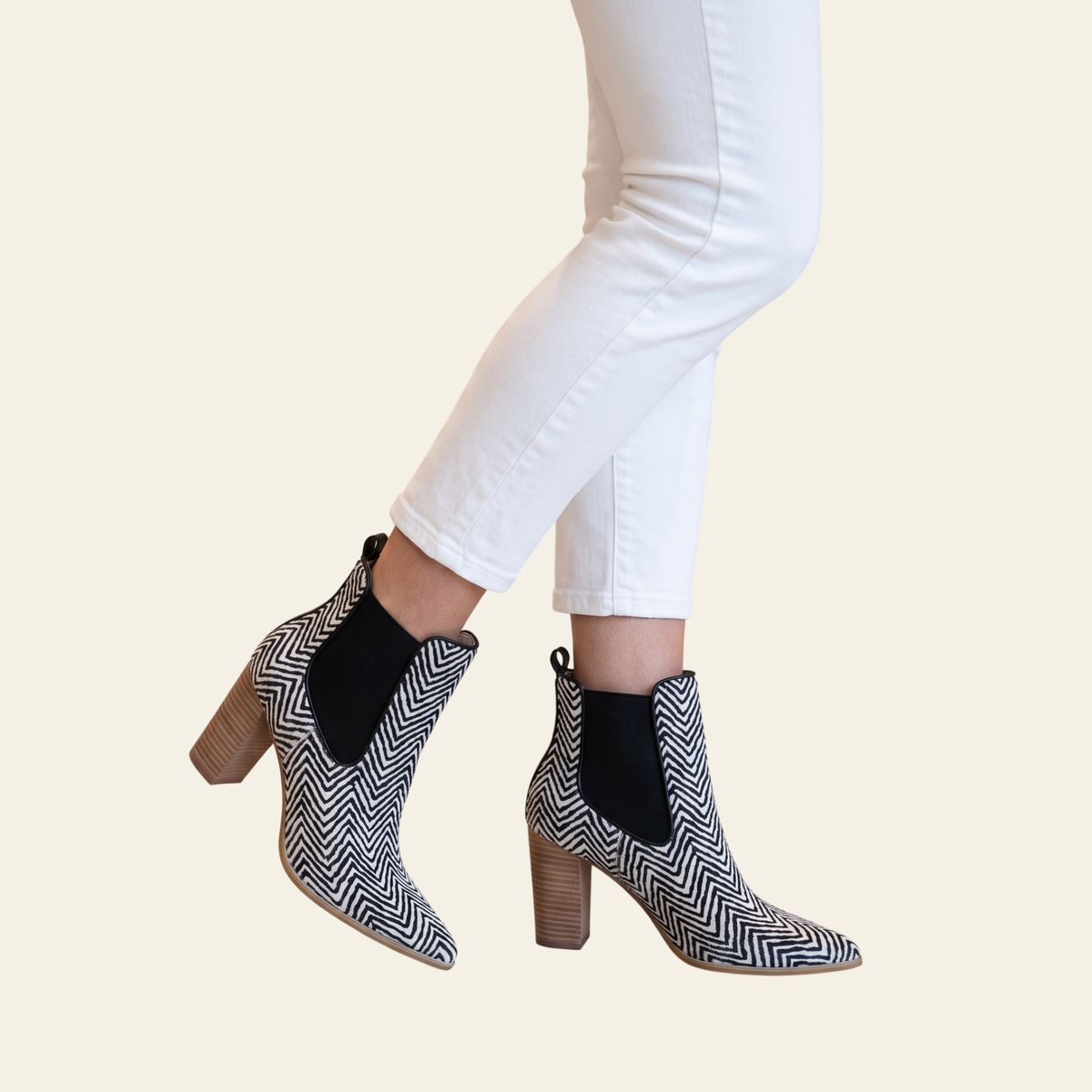 A person wearing black and white herringbone-patterned ankle boots with black elastic side panels and block heels, paired with white pants, against a solid light beige background.