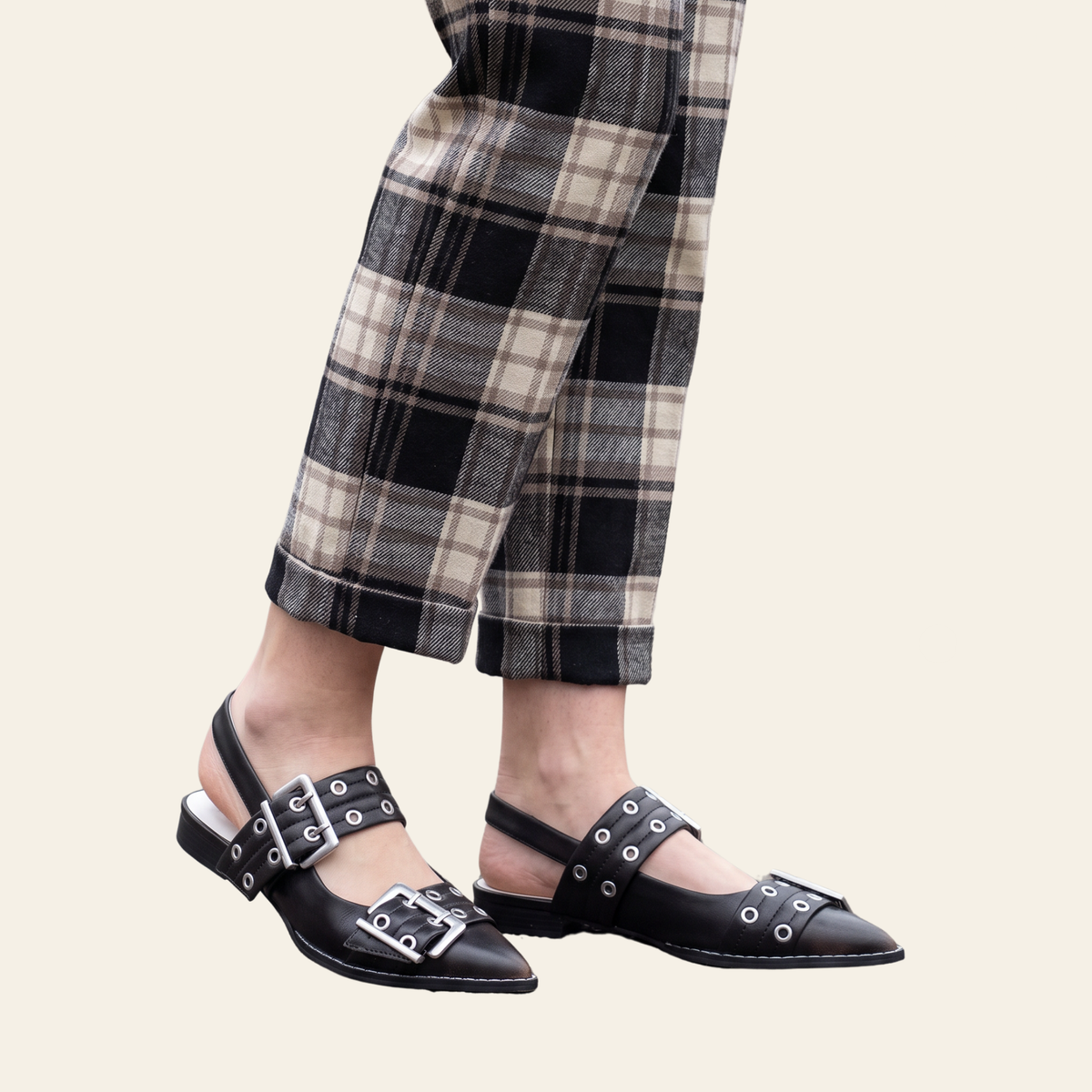 Brown pointed-toe slingback flats with silver buckle and grommet details, shown on a model wearing beige and black plaid trousers against a light cream background.