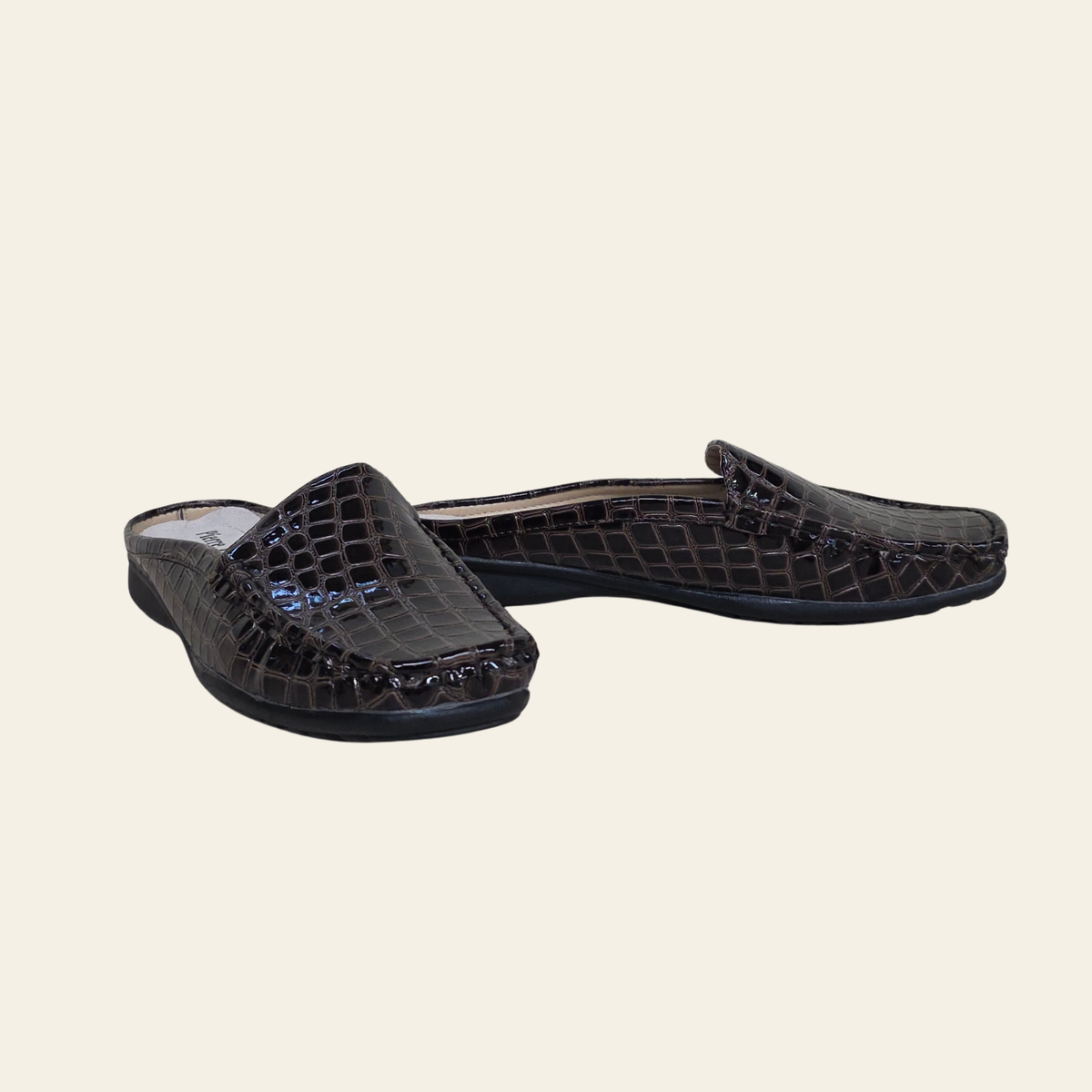A pair of dark brown crocodile-embossed slip-on mules with a glossy finish, displayed on a solid light beige background.