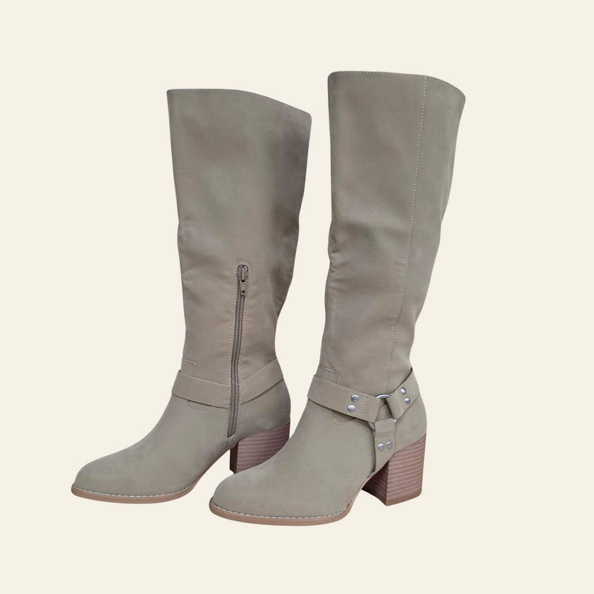A pair of knee-high taupe suede boots with side zippers and buckle strap details, shown against a solid cream background.