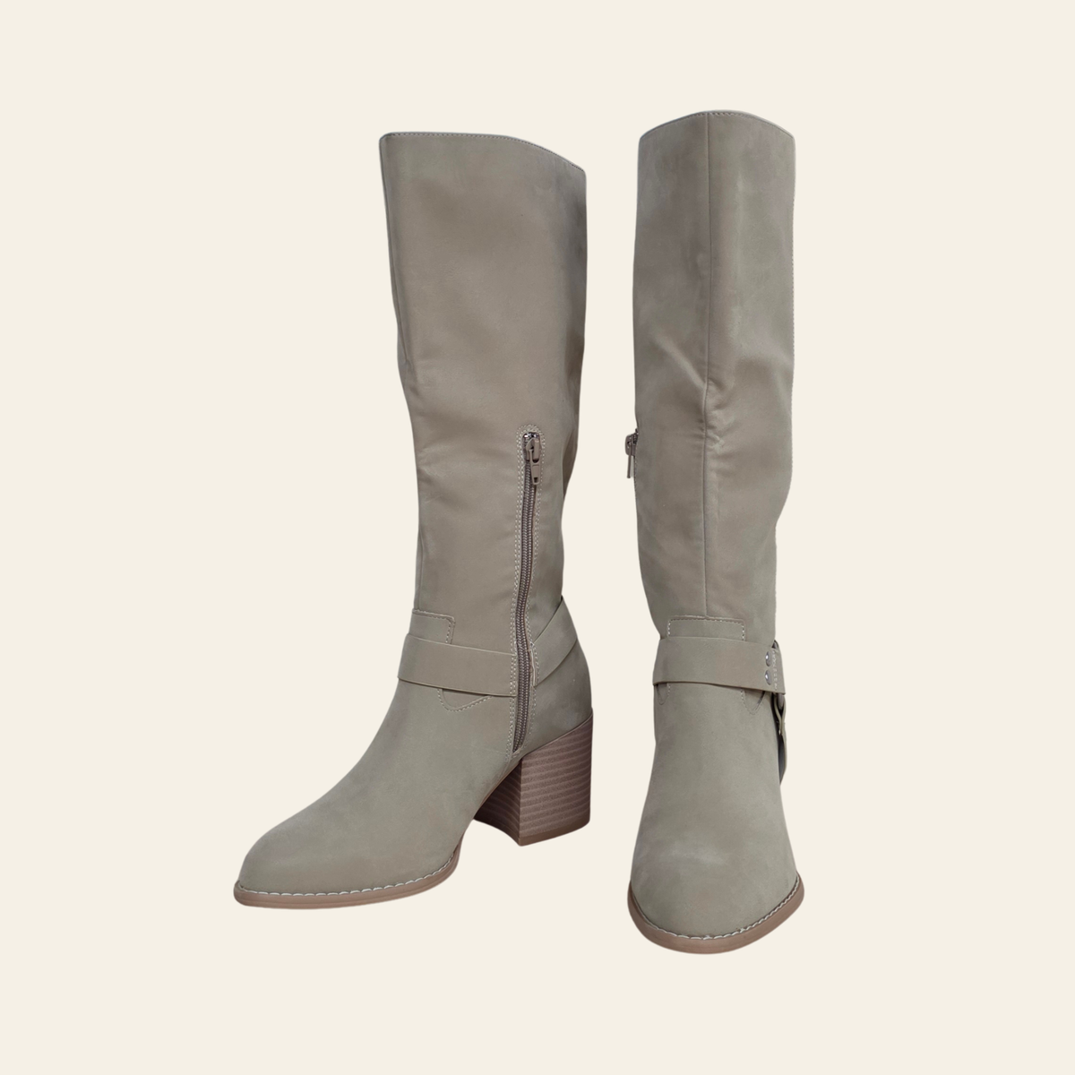 A pair of knee-high taupe suede boots with side zippers and buckle straps, shown against a solid cream background.