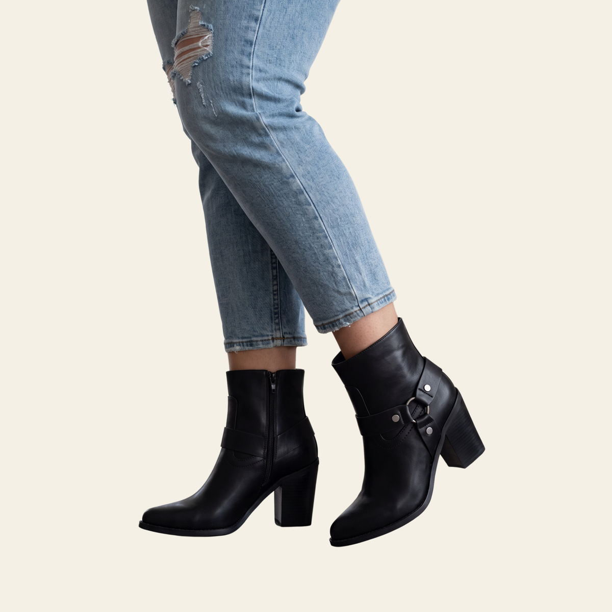 Black leather ankle boots with block heels and side buckle details, shown on a model wearing light blue ripped jeans against a solid cream background.
