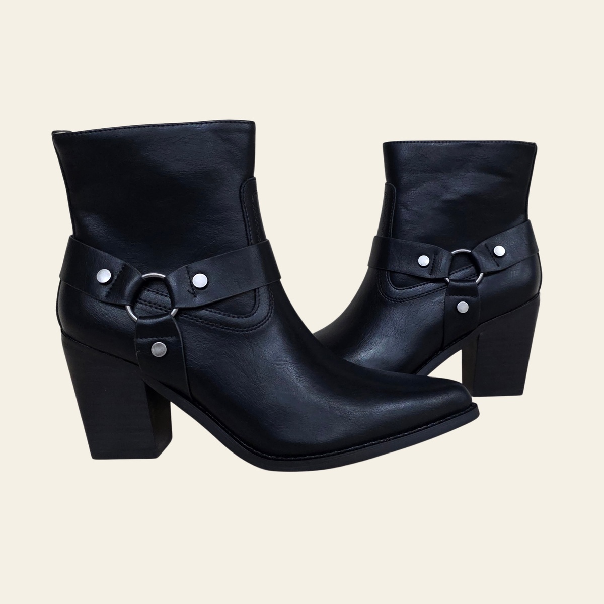 A pair of black leather ankle boots with block heels, pointed toes, and decorative strap details with silver studs and rings, shown against a solid light beige background.