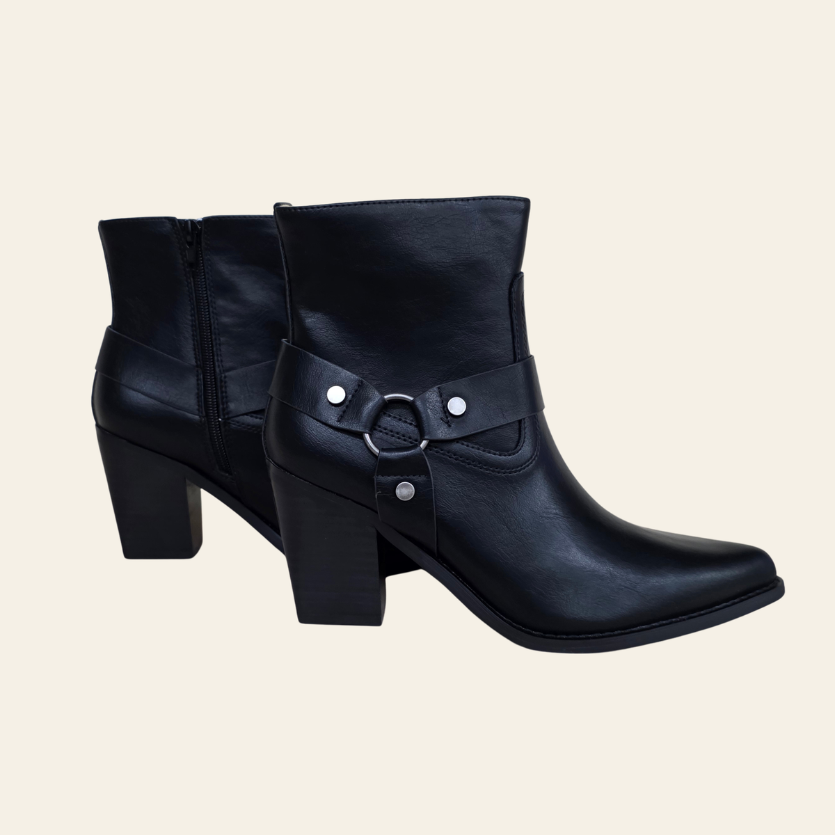 A pair of black faux-leather ankle boots with a block heel, pointed toe, and a decorative strap with silver studs and a ring detail on the side, shown against a solid light beige background.