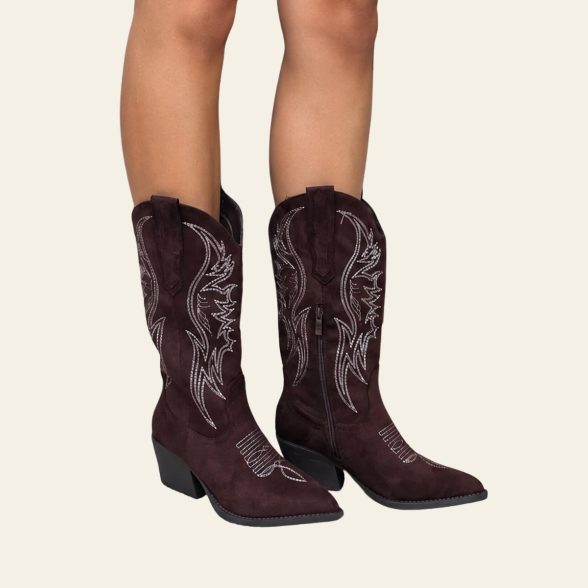 A pair of dark burgundy western cowboy boots with white embroidered stitching, shown on a model's feet against a light beige background.