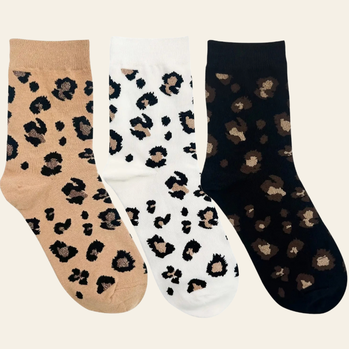 Three pairs of ankle socks with a leopard print pattern, shown side-by-side on a light cream background. The left sock is beige with black and brown spots, the middle sock is white with black and brown spots, and the right sock is black with brown and beige spots.