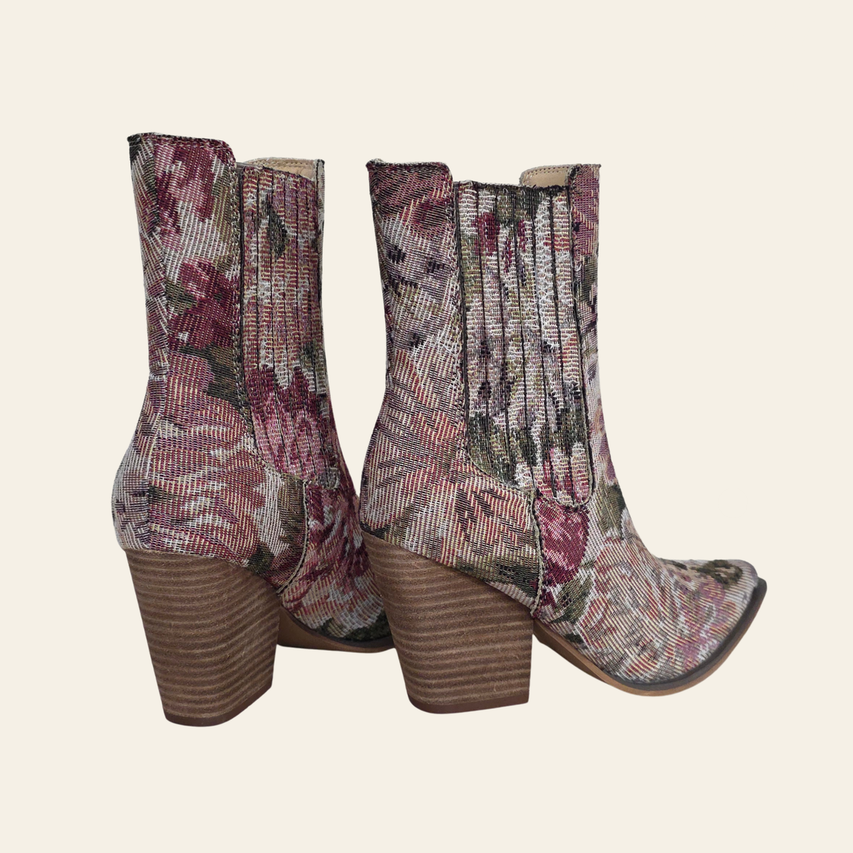 A pair of ankle boots with a floral tapestry pattern in dusty pink, burgundy, and green, featuring a brown stacked block heel and a pointed toe, shown against a solid cream background.