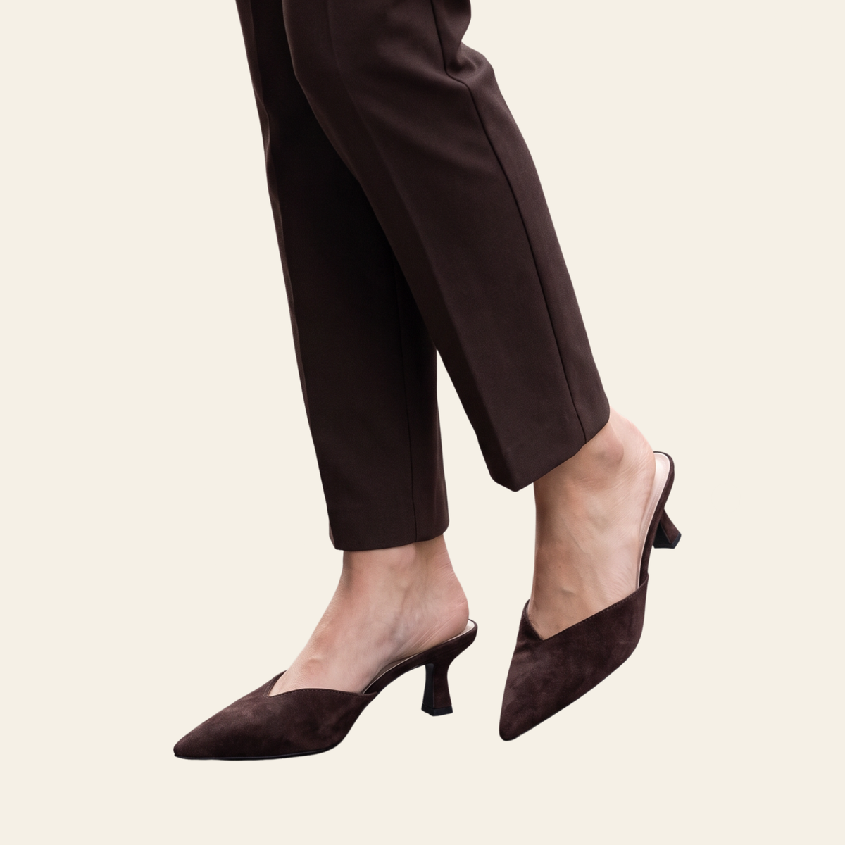 A person wearing dark brown pointed-toe mule heels with a low block heel and matching dark brown trousers, shown against a plain light beige background.