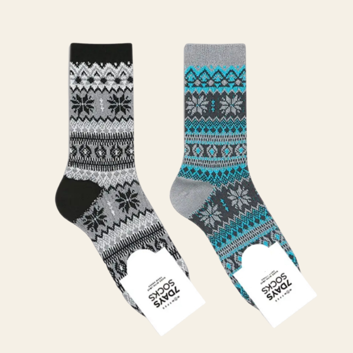 Two pairs of patterned crew socks displayed on a light beige background. One pair is black, gray, and white with a Fair Isle snowflake pattern, and the other is gray, blue, and black with a similar pattern.