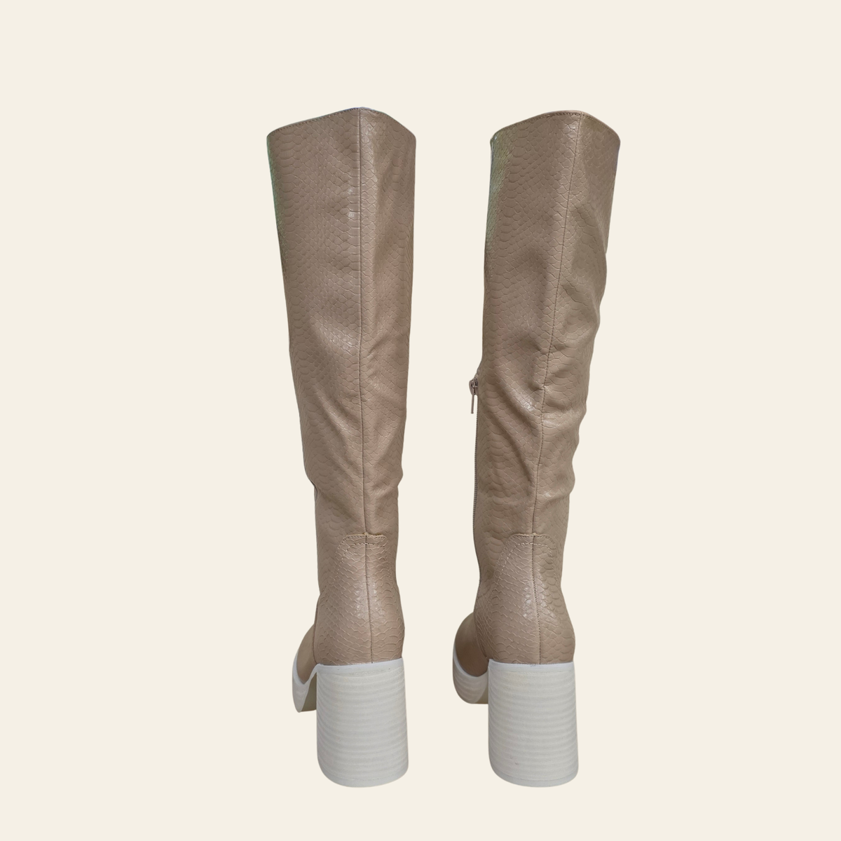 A pair of knee-high boots in a textured taupe-beige faux leather, shown from the back with chunky white block heels, set against a solid cream background.