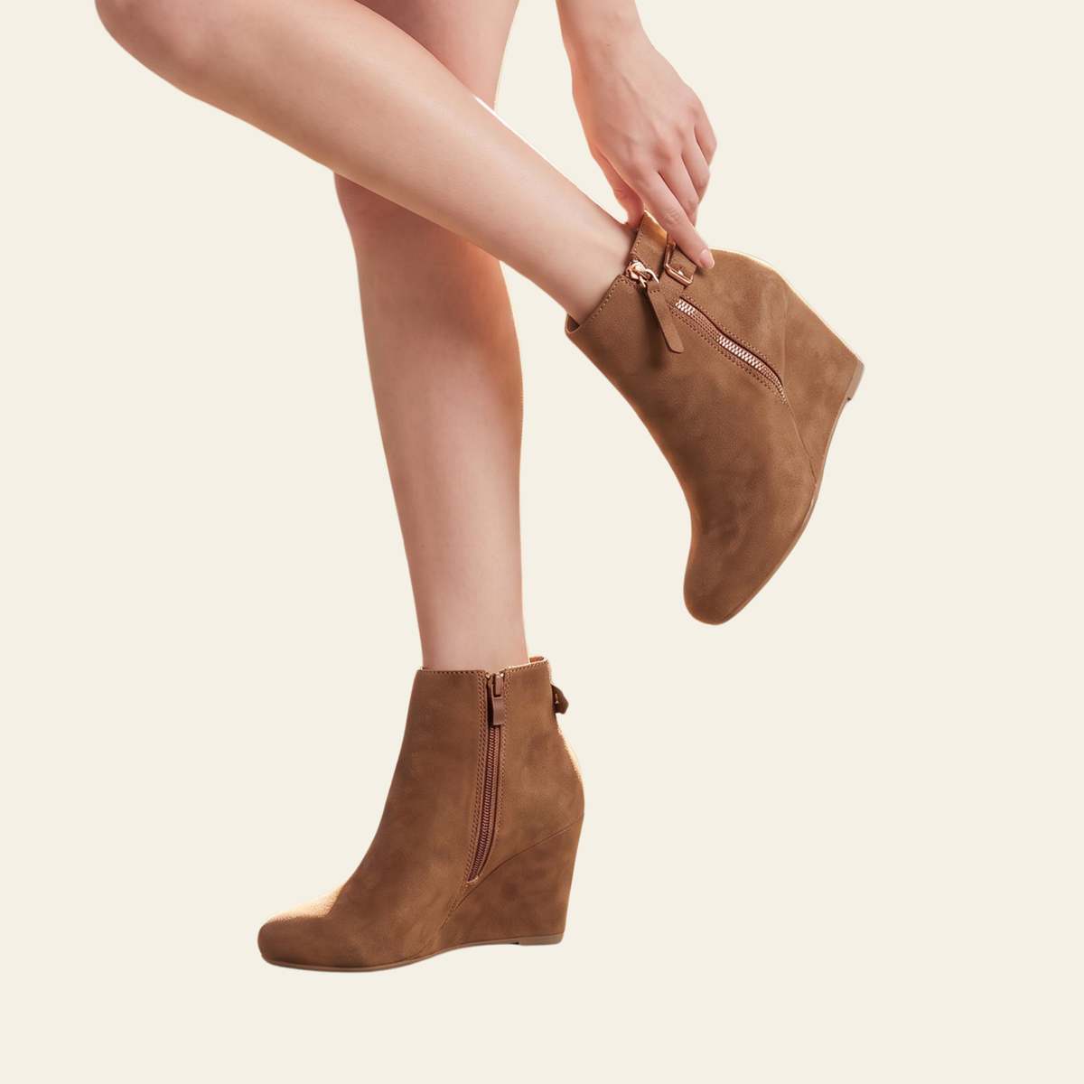A person wearing tan suede wedge ankle boots with side zippers, shown against a plain light beige background. One boot is on the foot and the other is being adjusted by a hand.
