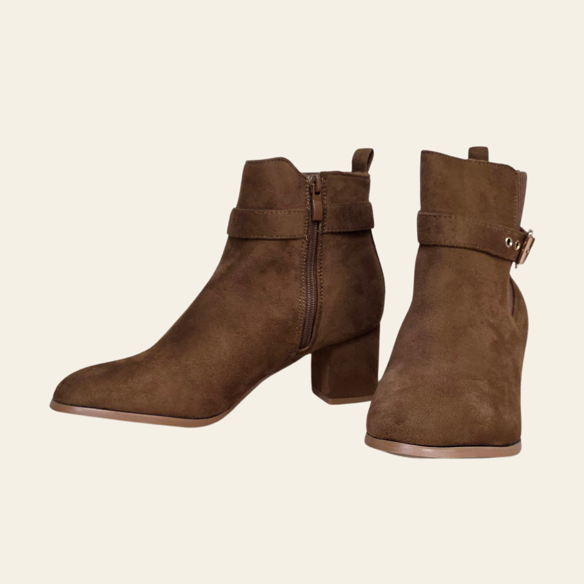 A pair of brown suede ankle boots with block heels, side zippers, and decorative straps with metal buckles, shown against a solid light beige background.