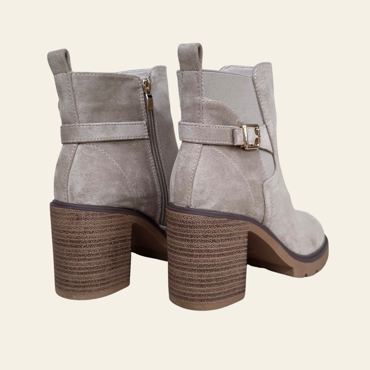 A pair of taupe suede ankle boots with block heels, side zippers, and decorative buckle straps, shown from a three-quarters angle against a light cream background.