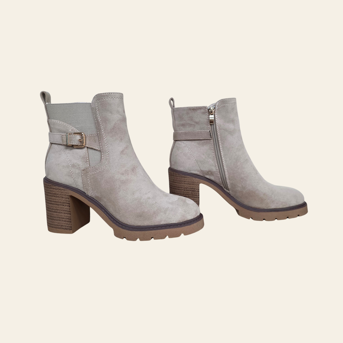 A pair of taupe suede ankle boots with brown block heels, a side buckle strap on one boot, and a side zipper on the other, shown against a solid cream background.