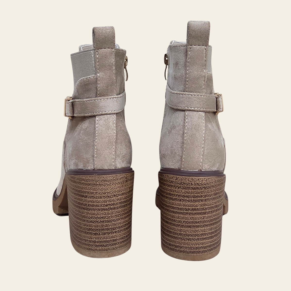 A pair of taupe suede ankle boots with block heels and side buckle straps, shown from the back against a light cream background.