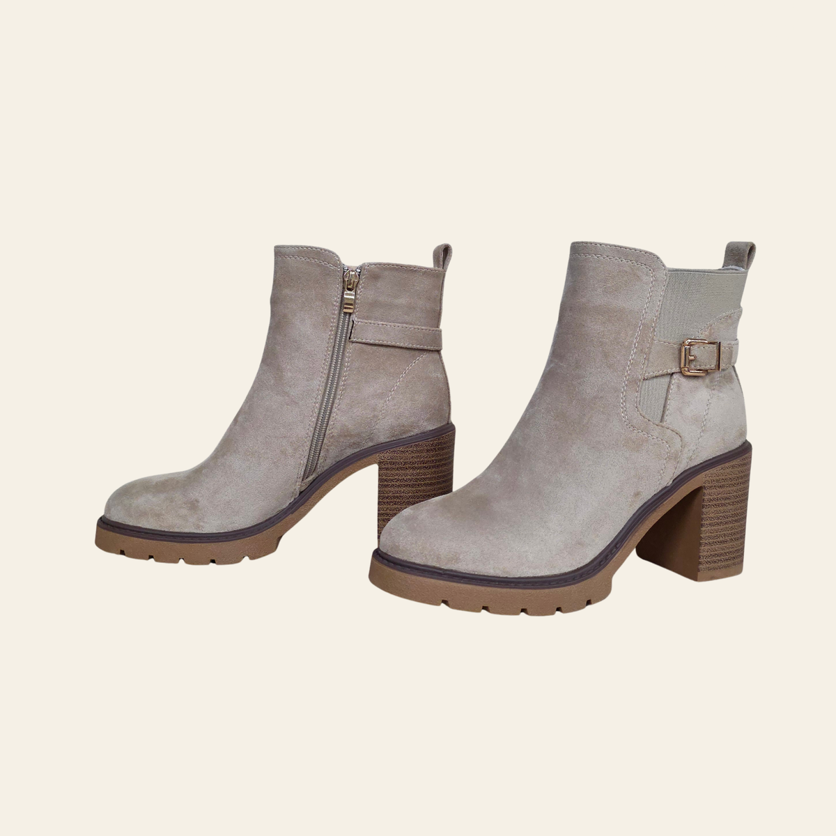 A pair of taupe suede ankle boots with block heels, featuring a side zipper on one boot and a decorative buckle strap on the other, set against a solid cream background.