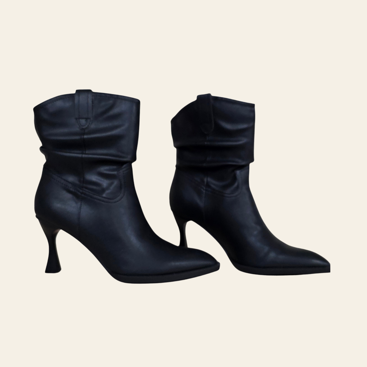 A pair of black leather ankle boots with a pointed toe and a unique curved kitten heel, shown against a solid light beige background.