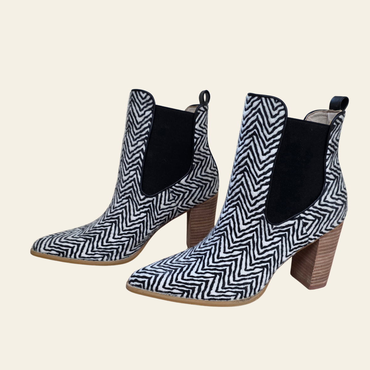A pair of women's ankle boots featuring a black and white herringbone pattern on the toe and heel, with black elastic side panels and stacked brown block heels, shown against a solid light beige background.