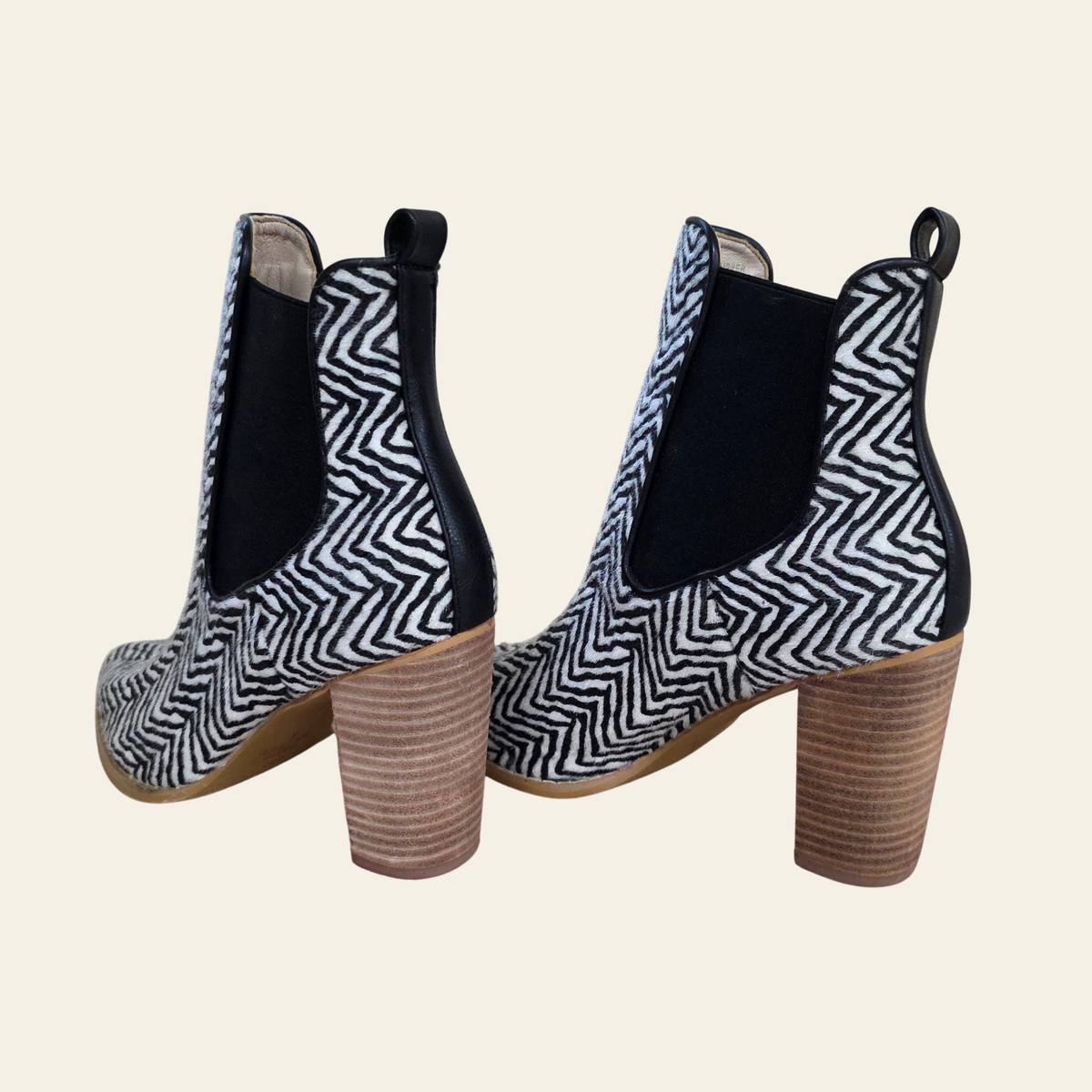A pair of women's ankle boots with a black and white chevron pattern on the toe caps, black elastic side panels, and stacked brown block heels, shown against a solid light beige background.