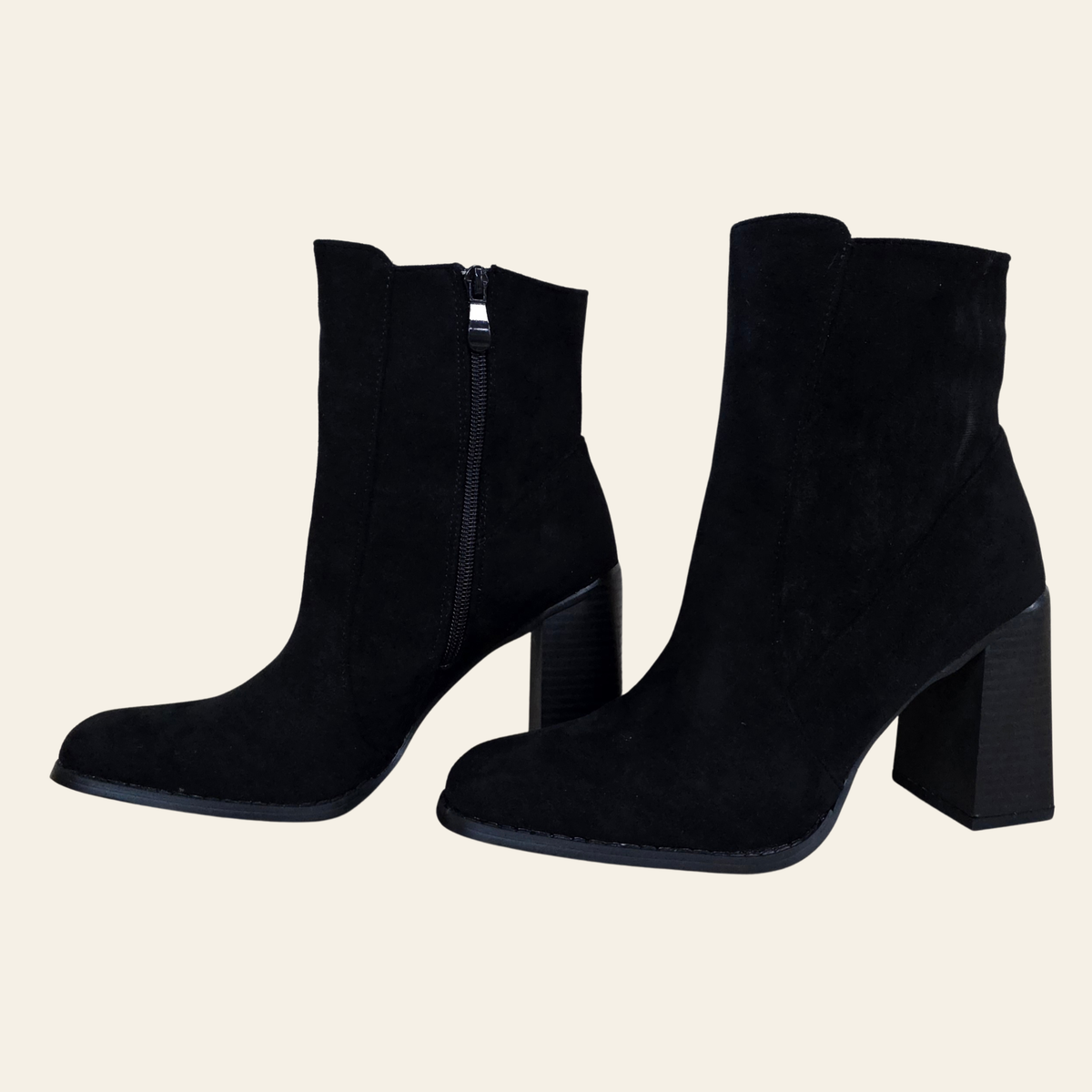 A pair of black suede ankle boots with block heels and side zippers, displayed against a solid light beige background.