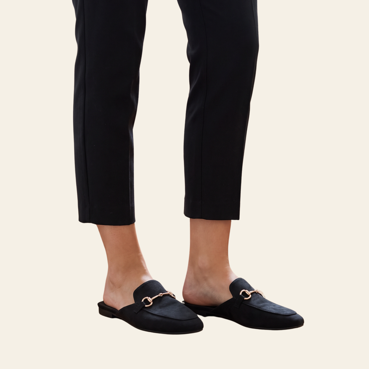 Black suede mule loafers with a gold-tone horsebit detail, shown on a model wearing black cropped trousers against a light cream background.