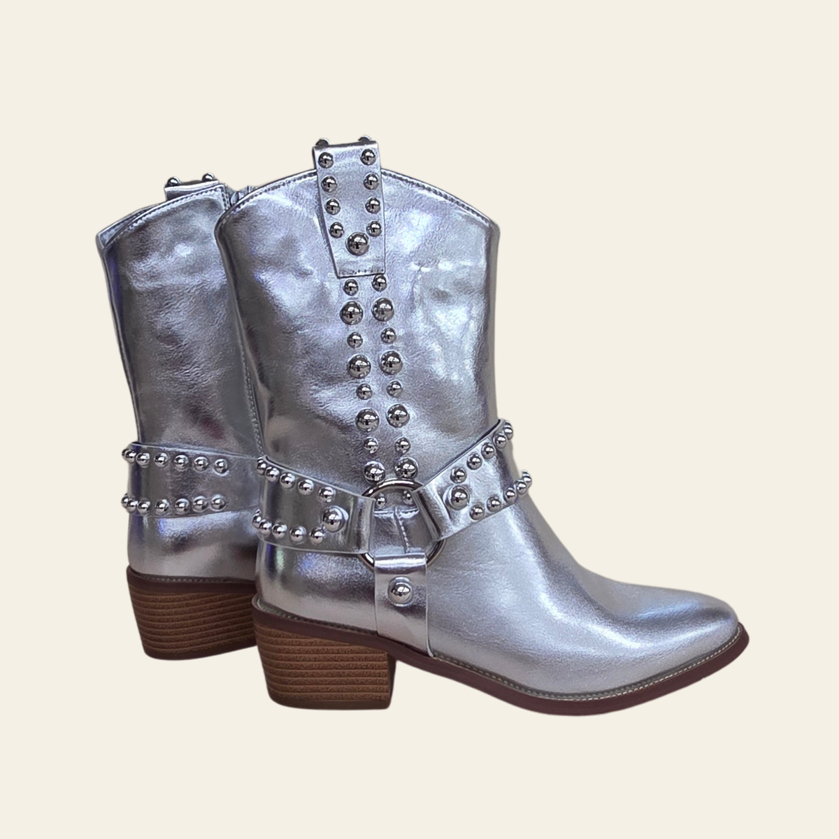 A pair of metallic silver western-style boots with brown stacked heels and silver studded straps, shown against a solid light beige background.