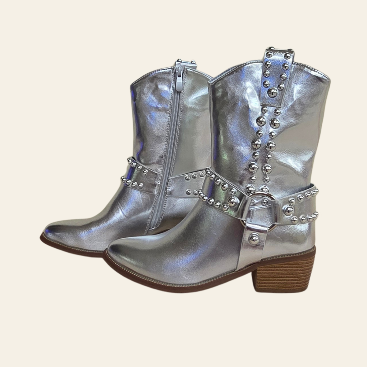 A pair of metallic silver western-style boots with studded straps, side zippers, and brown stacked block heels, shown against a solid light beige background.