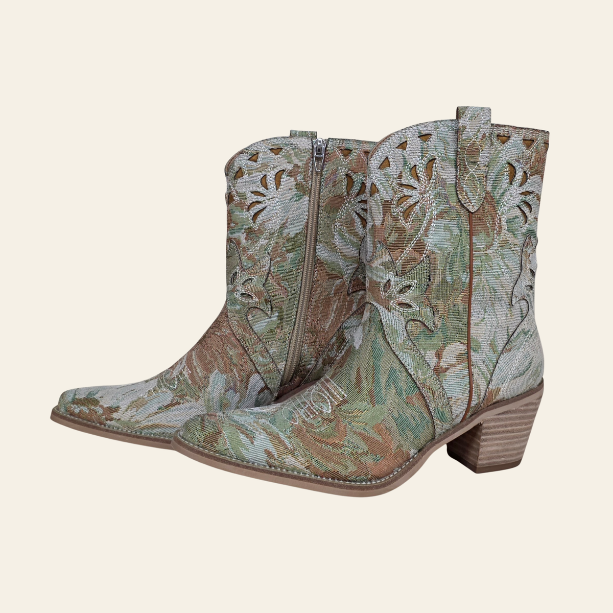 The Amaryllis - western ankle boots with a floral woven pattern in sage green, brown, and beige, featuring a pointed toe and stacked wood-look heels, set against a solid cream background.