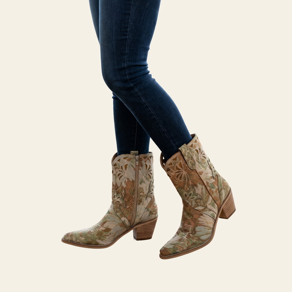 A pair of women's western-style ankle boots with a beige, brown, and green floral pattern, shown on a model wearing dark blue skinny jeans against a light cream background.