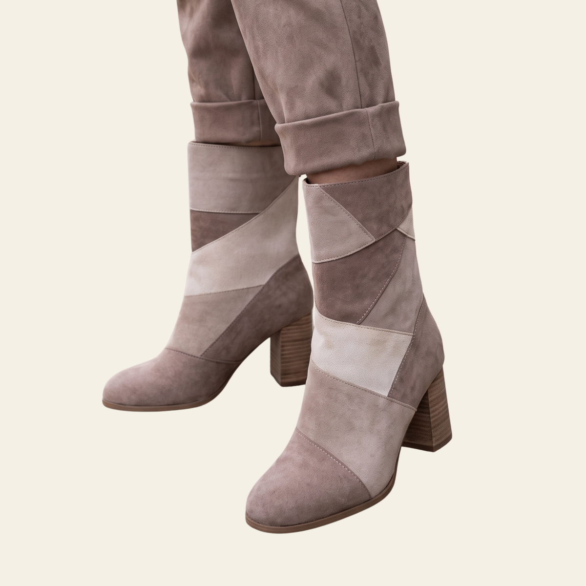 A pair of women's ankle boots in taupe and beige suede with geometric paneling and block heels, shown on a model wearing cuffed trousers against a solid cream background.