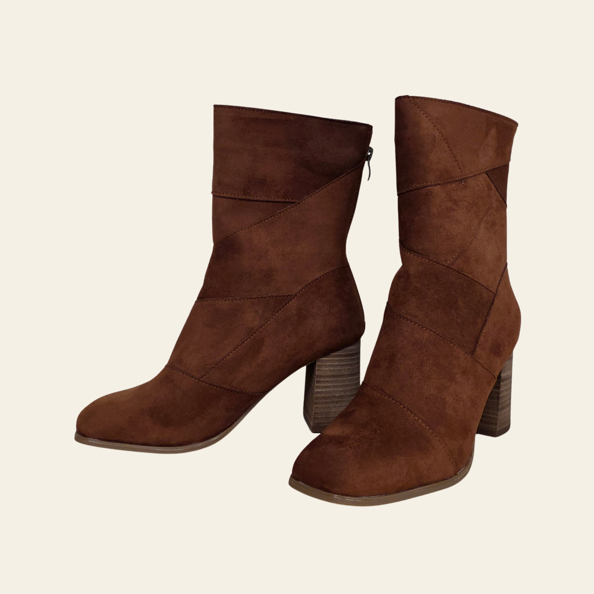 A pair of brown suede ankle boots with geometric paneling and stacked block heels, shown against a solid off-white background.